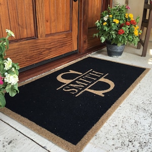 May include: A black doormat with a tan border and the text "Smith" in a split monogram design. The monogram is in tan and features a stylized letter "S".