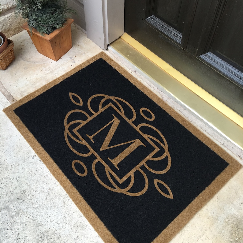 May include: A black doormat with a tan border and a gold monogram "M" in a decorative frame.