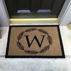 May include: A brown doormat with a black border and a black wreath with the letter "W" in the center.