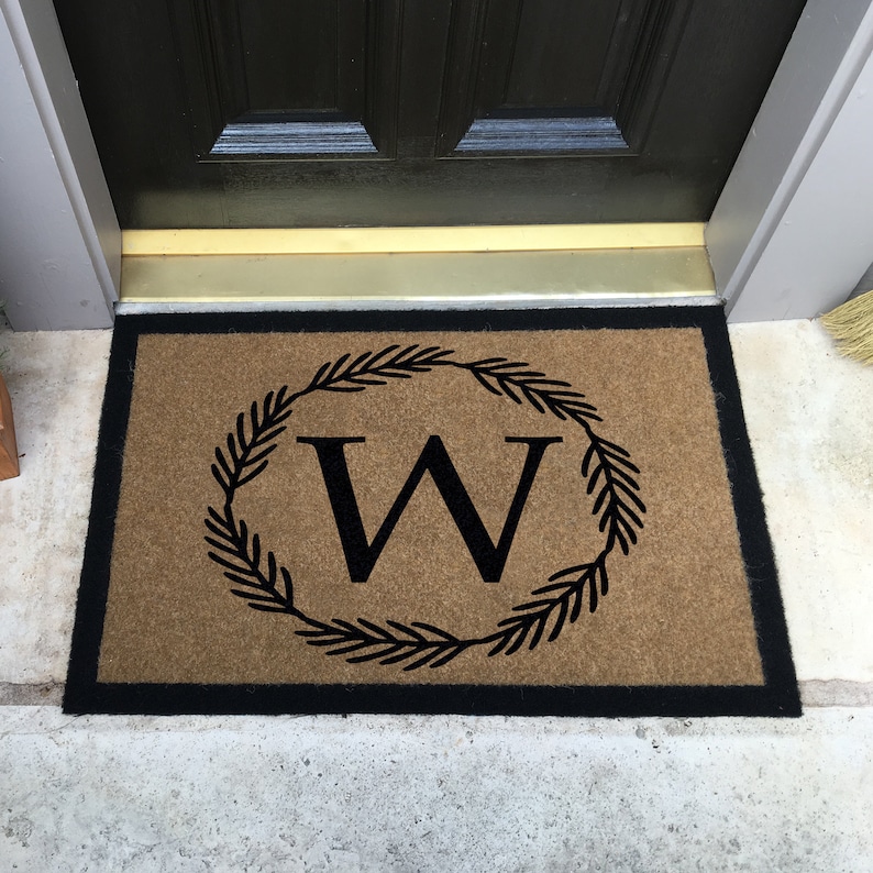 May include: A brown doormat with a black border and a black wreath with the letter "W" in the center.