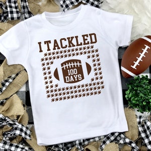 K&ouml;nnte beinhalten: Wei&szlig;es T-Shirt mit braunem Text, der "I Tackled 100 Days" lautet. Der Text ist von einem braunen Umriss eines Fu&szlig;ballfeldes mit braunen Fu&szlig;b&auml;llen innerhalb des Umrisses umgeben. Ein brauner Fu&szlig;ball liegt auf einer schwarz-wei&szlig; karierten Oberfl&auml;che.