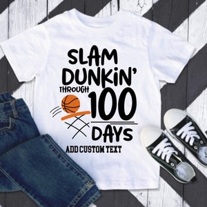 May include: White t-shirt with the text "SLAM DUNKIN' THROUGH 100 DAYS" and a basketball graphic. Includes the text "ADD CUSTOM TEXT". The shirt is paired with blue jeans and black sneakers.