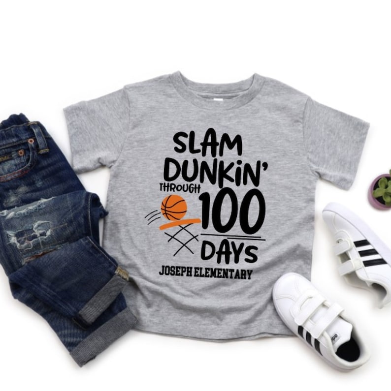 May include: A gray t-shirt with the text "SLAM DUNKIN' THROUGH 100 DAYS" and a basketball graphic. The shirt also includes "JOSEPH ELEMENTARY" at the bottom. The shirt is paired with blue jeans and white sneakers.