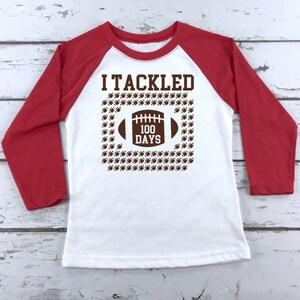 K&ouml;nnte beinhalten: Wei&szlig;es und rotes Langarm-Baseball-T-Shirt mit dem Text "I Tackled 100 Days" und einer braunen Football-Grafik.