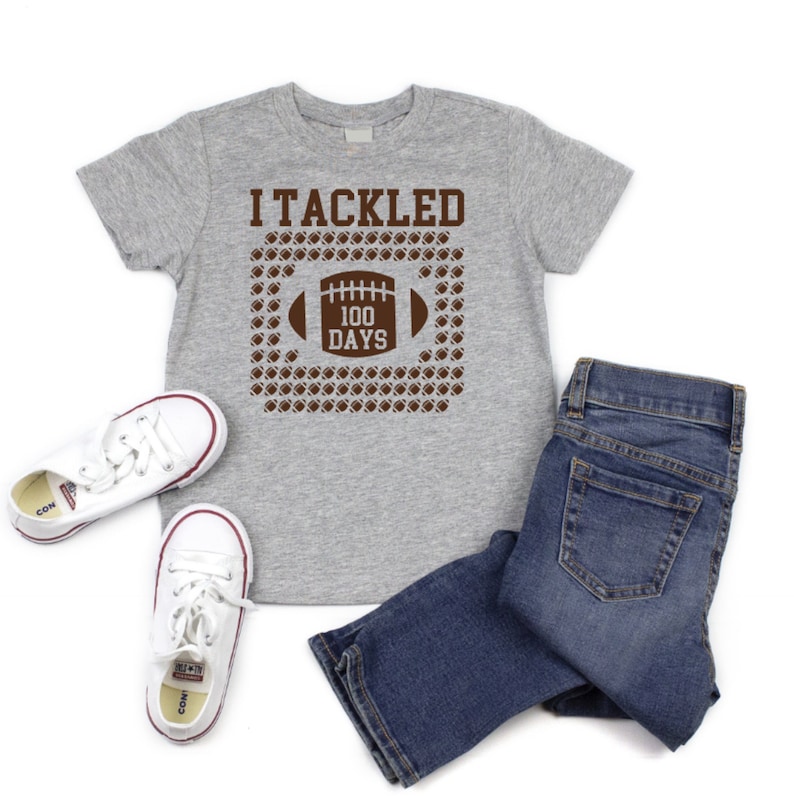 K&ouml;nnte beinhalten: Ein graues T-Shirt mit einer braunen Grafik eines Fu&szlig;balls und dem Text "I Tackled 100 Days".