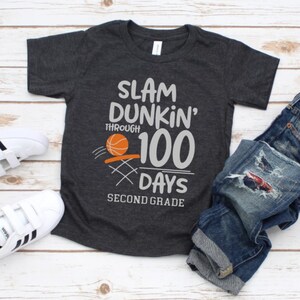 May include: Dark gray t-shirt with the text "SLAM DUNKIN' THROUGH 100 DAYS SECOND GRADE" and a basketball graphic. The shirt is paired with blue denim jeans and white sneakers with black stripes.