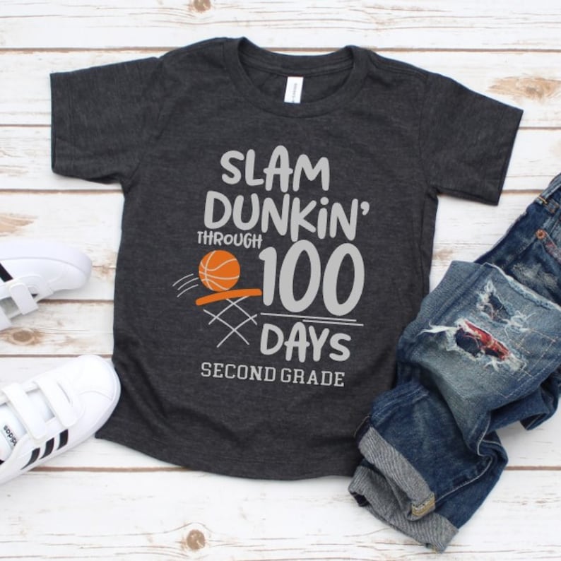 May include: Dark gray t-shirt with the text "SLAM DUNKIN' THROUGH 100 DAYS SECOND GRADE" and a basketball graphic. The shirt is paired with blue denim jeans and white sneakers with black stripes.