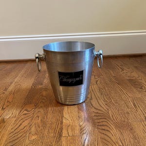 May include: A silver-toned metal champagne bucket featuring a black rectangular plaque on the front with the word Champagner written in white script. The bucket has two side handles with circular rings and horizontal grooved detailing.
