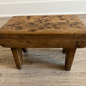 May include: A rustic wooden stool with a rectangular top and three legs. The stool is made of weathered wood and has a natural brown color.