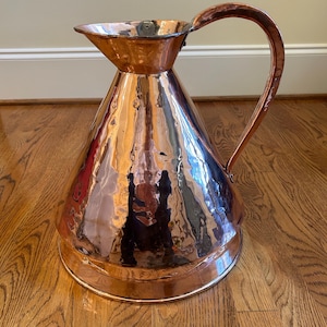 May include: A large, copper pitcher with a hammered finish and a wide, flared spout. The pitcher has a single handle and a rounded base.