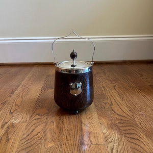 May include: A dark brown wooden ice bucket with a polished silver-tone metal lid and a matching swing handle. The bucket features a small, shield-shaped silver-tone metal plaque on the front and a round wooden knob on the center of the lid.