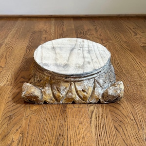 May include: A circular wooden pedestal base featuring a weathered, light-colored top surface and a decorative, carved lower section with a distressed gold and brown finish. The base has a square-like footprint with ornate, sculpted corner details.