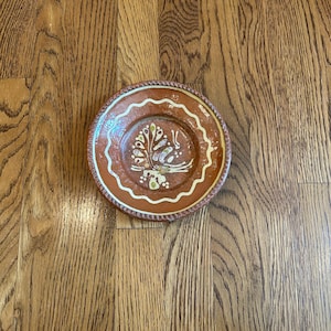 May include: A round, terracotta-colored plate with a decorative tree design in the center. The plate has a white wavy border and a rope-like edge. The tree design features white, yellow, and green accents. The plate is set on a wooden surface.