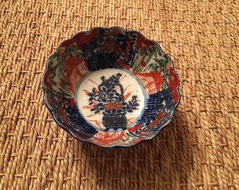 May include: A small, round ceramic bowl with a scalloped edge. The interior features a central dark blue floral motif surrounded by panels of orange, green, and blue patterns. The white base contrasts with the intricate, multi-colored hand-painted designs.