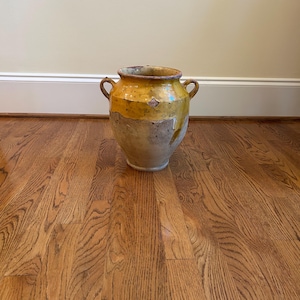 May include: A large, antique-style ceramic jar with a wide body and two small handles. The jar features a two-tone design, with a yellow-ochre upper section and a lighter, mottled lower section. The jar sits on a hardwood floor.