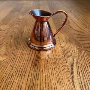 May include: A small copper pitcher with a rounded base and a handle. The pitcher has a shiny, metallic finish.