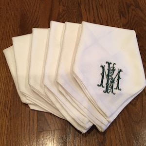 May include: A stack of white cloth napkins with green embroidered initials "NM" in a decorative font.
