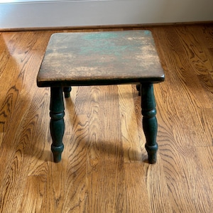 May include: A small, square wooden step stool with a distressed, aged appearance. The top surface is a faded green and brown, while the legs are a darker green. The stool is likely vintage or antique, with a rustic aesthetic.