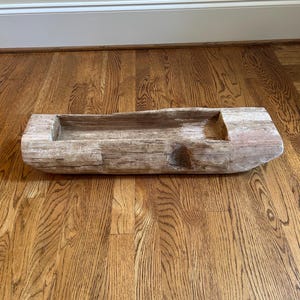 May include: A long, rectangular wooden dough bowl with a weathered, light brown finish. The object features a hollowed-out center, rough textured exterior surfaces, and visible wood grain patterns throughout its elongated, sturdy construction.