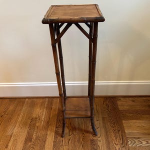 May include: A tall, dark brown plant stand constructed from bamboo-style wood. It features a square top surface, four slender legs connected by angled support braces, and a smaller square shelf positioned near the base of the structure.