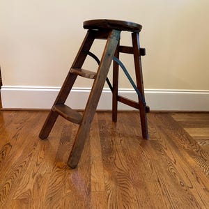 May include: A vintage wooden step stool with a dark brown finish. The stool has a round seat and two steps, with metal supports. The step stool is designed to fold for storage. The stool is on a hardwood floor.