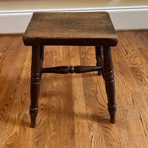 May include: A dark brown wooden stool with a square seat and four turned legs connected by horizontal stretchers. The wood features a visible grain pattern and a worn finish, showing signs of age and use on the surface and edges of the seat.