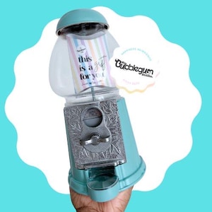 Puede incluir: Una máquina de chicles turquesa con una ranura para monedas plateada y un tarro de vidrio transparente. La máquina tiene un cartel que dice "This is a gift for you".