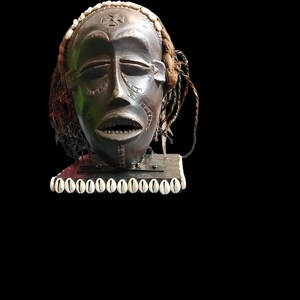 May include: A dark brown wooden mask with intricate carvings, adorned with white shells and braided hair. The mask features closed eyes, an open mouth, and detailed facial patterns, mounted on a rectangular base.