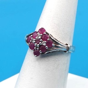 May include: A silver ring featuring a cluster of round, deep pinkish-red rubies arranged in a diamond-shaped setting. Small, clear accent stones are interspersed between the rubies. The band has a split-shank design that tapers toward the back.