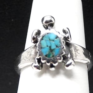May include: A silver ring featuring a turtle-shaped centerpiece. The turtle shell is inlaid with small, irregular pieces of bright blue turquoise. The ring band is smooth and polished, with the turtle head, four legs, and tail extending outward.