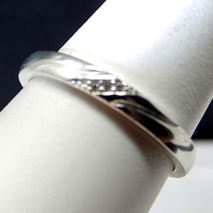 May include: A polished silver-toned ring featuring a smooth, rounded band with a subtle, diagonal channel set with three small, clear, sparkling stones. The metal surface reflects light, highlighting the curved design and the embedded stones.