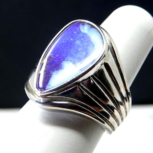 May include: A silver ring with a pear-shaped opal stone. The opal displays a mix of blue, purple, and white colors. The ring's band has a layered design, adding to its unique appearance. The ring is shown on a white surface.