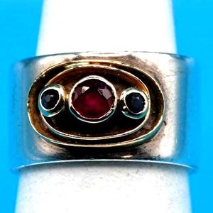 May include: A wide silver band ring featuring a central oval gold setting. The gold setting contains one round faceted red ruby in the center, flanked by two smaller round dark blue sapphires, all set in a horizontal arrangement.