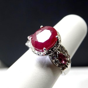 May include: A silver ring featuring a large, oval-cut ruby gemstone as the centerpiece. The ruby is a deep red color and is surrounded by smaller, clear gemstones. Additional gemstones adorn the ring's band, creating a sparkling effect.