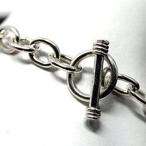 May include: A silver-toned chain link bracelet featuring a toggle clasp closure. The bracelet is composed of polished, rounded metal links connected to a circular ring and a perpendicular bar component that secures the jewelry piece together.