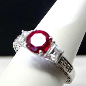 Può includere: Un anello d'argento con una grande gemma rossa scura rotonda come elemento centrale. Due gemme chiare quadrate più piccole affiancano la pietra principale. La fascia dell'anello presenta dettagli intricati.