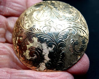 May include: A gold-colored, round compact with an ornate floral design. The compact has a textured surface with detailed engravings of leaves and swirling patterns. The edges of the compact are also decorated with a delicate, repeating pattern.