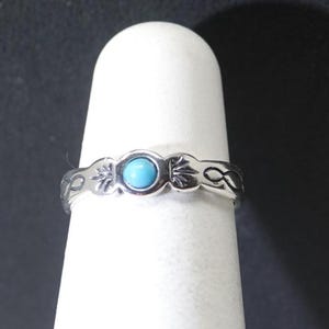 May include: A silver ring featuring a central round turquoise cabochon stone. The band is crafted from polished silver and displays engraved decorative patterns on both sides of the stone, including leaf-like motifs and infinity-style symbols.