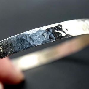 May include: A silver-colored metal bangle bracelet with a hammered, textured surface. The band features a reflective, metallic finish with irregular indentations across the exterior, creating a rugged, uneven appearance along the entire circumference.