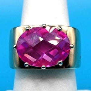 May include: A wide gold band ring featuring a large, oval-cut, vibrant pink gemstone set in the center. The gemstone is secured by six small metal prongs, and the band has a smooth, polished finish that wraps around the finger.