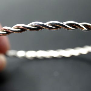 May include: A polished silver-toned bangle bracelet featuring a continuous twisted rope design. The metal surface reflects light, highlighting the spiral texture of the circular band. The bracelet has a uniform thickness and a smooth, metallic finish.
