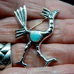 May include: A silver-toned roadrunner brooch with a turquoise stone set in the center. The brooch is detailed with feathers and a running pose, and has a pin on the back.