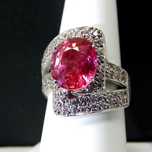 May include: A silver ring featuring a large, oval-cut, pink gemstone as the centerpiece. The ring is surrounded by smaller, light purple gemstones. The band has a split design, with additional small gemstones. The ring is displayed on a white stand.