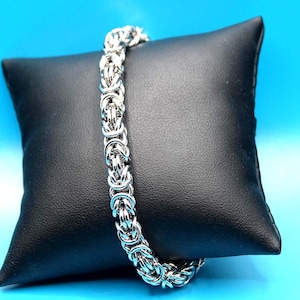 May include: A silver-toned chain bracelet featuring a complex Byzantine link pattern. The bracelet is constructed from numerous interlocking metal rings that create a dense, cylindrical, and textured design with a polished, reflective finish.