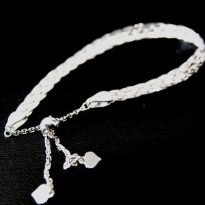 May include: A silver-colored bracelet with a woven design. The bracelet has a sliding clasp with two heart-shaped charms dangling from the end. The bracelet is displayed against a black background.