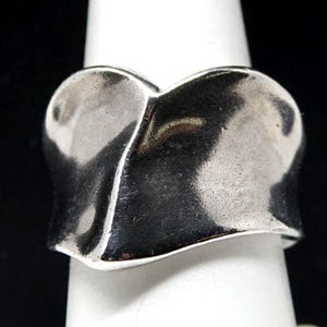 May include: A wide silver ring with a polished, reflective surface and a distinct, undulating wave-like shape. The metal band features smooth, organic curves that create a continuous, flowing silhouette around the finger, showcasing a bright metallic finish.