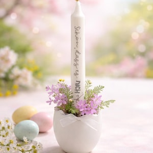 Może przedstawiać: Biała świeca z napisem "Schön, dass es dich gibt" umieszczona w białym, jajowatym świeczniku wypełnionym fioletowymi kwiatami i zielenią. Pastelowe pisanki i białe kwiaty w tle.