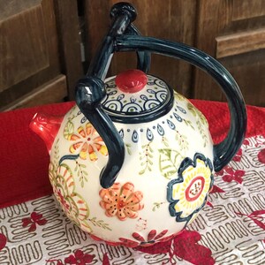 May include: A white ceramic teapot with a floral design. The teapot features a dark blue handle and spout, and a red lid. The floral pattern includes orange, yellow, and green flowers and leaves. The teapot is sitting on a red surface.