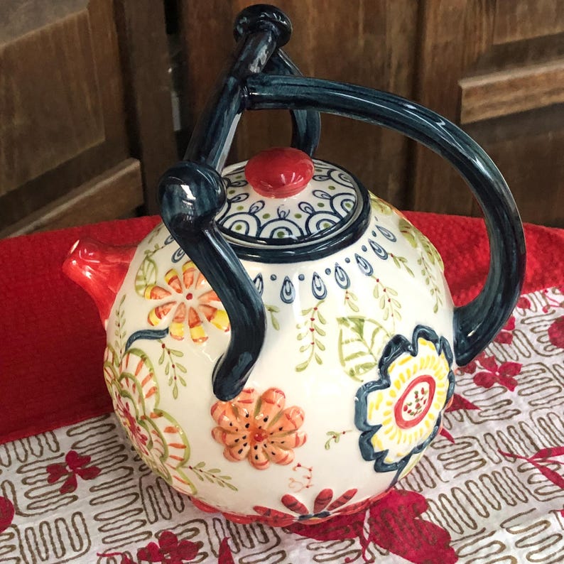 May include: A white ceramic teapot with a floral design. The teapot features a dark blue handle and spout, and a red lid. The floral pattern includes orange, yellow, and green flowers and leaves. The teapot is sitting on a red surface.