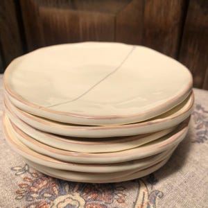 May include: A stack of five round ceramic plates with an off-white, glossy finish. Each plate features an irregular, organic edge with a thin, light brown painted rim. A single, faint, diagonal line runs across the surface of the top plate.